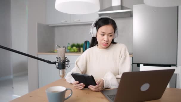 Aziatische vrouw streamer maakt een live podcast voor sociale netwerken, op afstand werken tijdens het zitten in de keuken, met behulp van laptop en tablet, werken in de context van de coronavirus pandemie, quarantaine. — Stockvideo