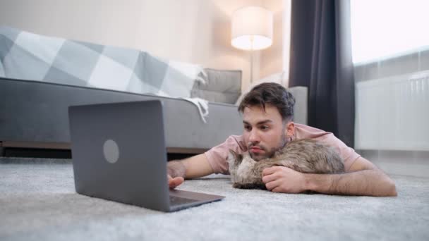 Un hombre joven con un gato yace en el suelo en la sala de estar y trabaja en una computadora portátil, autoaislamiento en casa durante la cuarentena, trabajo remoto. — Vídeo de stock