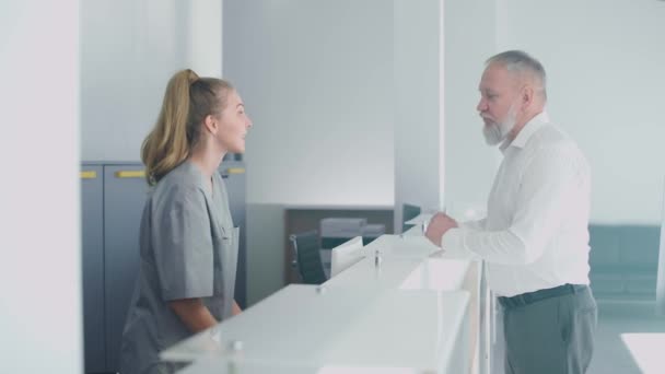 Nemocniční recepce, zdravotní sestra registruje staršího muže pacienta na schůzku s lékařem v moderní lékařské klinice, lidé v ochranných lékařských maskách, koronavirová pandemie. — Stock video