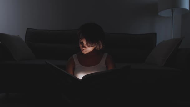 Portrait of young female sitting at home on the floor and reading a book, warm light, self-isolation during the pandemic, mystical lighting effect. — Stock Video