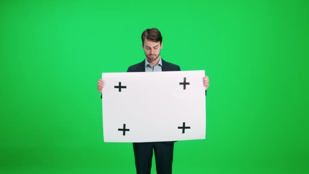 Mladý muž v obleku stojící a drží plakát z bílého papíru, šablona, seriózní pohled na fotoaparát na pozadí chroma klíč. — Stock video