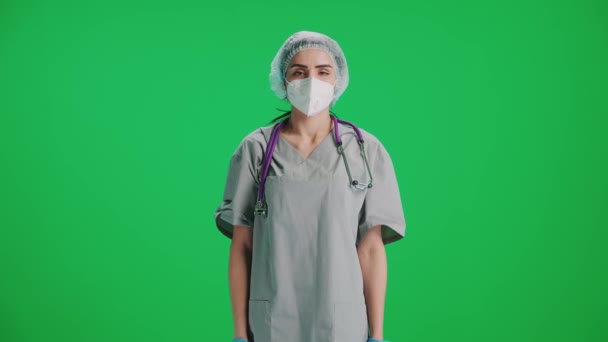 Retrato de jovem médica em equipamento de proteção e máscara médica, mulher olha para a câmera, proteção contra um covid-19, olhar sério para a câmera em um fundo de chave de croma.. — Vídeo de Stock