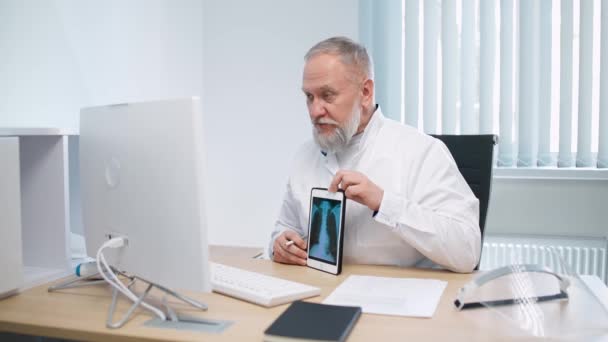 灰白头发的男医生坐在工作场所，通过视频通话与病人交流，医生在网上咨询病人，并在平板电脑屏幕上显示出病人的X光图像，这就是现代医院. — 图库视频影像