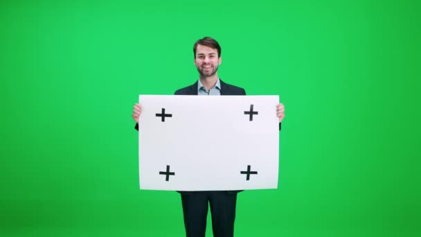 Mladý muž v obleku stojí a drží plakát z bílého papíru, šablony, usmívá se na fotoaparát na pozadí chroma klíče. — Stock video