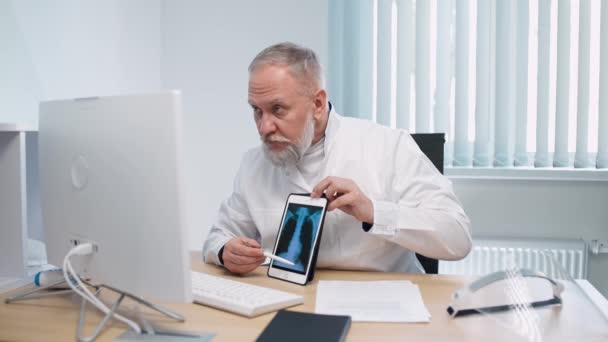 Il medico maschio dai capelli grigi siede sul posto di lavoro e comunica con il paziente tramite videochiamata, il medico consulta il paziente online, mostrando l'immagine a raggi X sullo schermo del tablet, moderno ospedale. — Video Stock
