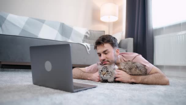 Junger Mann liegt mit Katze im Wohnzimmer auf dem Boden und arbeitet am Laptop, Selbstisolierung zu Hause während Quarantäne, Fernarbeit. — Stockvideo