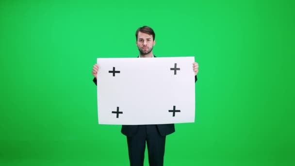 Mladý muž v obleku stojící a drží plakát z bílého papíru, šablona, seriózní pohled na fotoaparát na pozadí chroma klíč. — Stock video