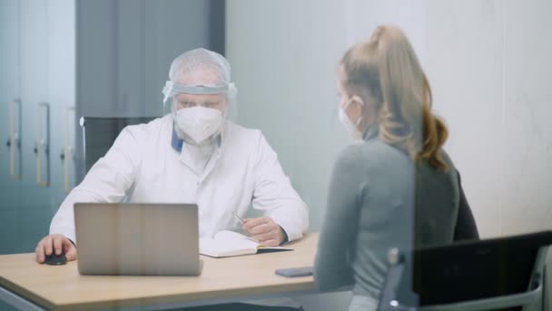 Moderne prive-kliniek, jonge vrouw bij een doktersafspraak, volwassen man arts en een jonge vrouw in beschermende medische maskers, een artsen bezoek tijdens de coronavirus pandemie, onderzoek door een — Stockvideo