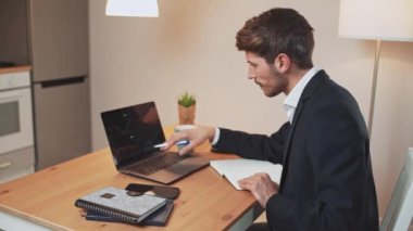 Takım elbiseli bir adam, evdeki uzak bir çalışmada mali analist, belgelerle ve finansal beyannamelerle çalışıyor, laptop ekranındaki bilgi grafiklerine ve belgelere bakıyor, bir salgın bağlamında uzaktan çalışma.