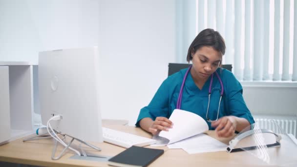 Ernstige vrouwelijke arts zit op het werk in een modern ziekenhuis en werkt met patiëntenonderzoeksdocumenten. — Stockvideo