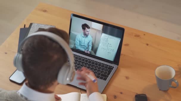 Hombre estudiante se sienta en un escritorio en casa y estudiar en línea utilizando una computadora portátil, estudiante aprende en una lección remota por videollamada con un profesor de hombres, la educación en las condiciones de la pandemia de coronovirus — Vídeo de stock