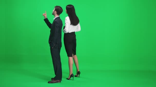 Man en vrouw in pakken staan in de groene ruimte van de chromakey, ze bespreken onderwerpen terwijl ze voor ze kijken, een sjabloon op een groene achtergrond, achteraanzicht. — Stockvideo