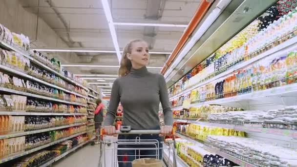 Mujer joven camina entre los estantes en una tienda de comestibles, visitando un supermercado, una mujer camina con un carrito de compras, 4k cámara lenta. — Vídeos de Stock
