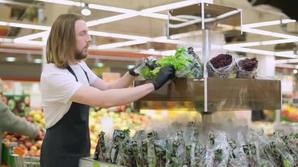 De mens vult producten aan op de planken, de werknemer legt de groene saladepakketten op de planken in een biologische winkel, werkt in een supermarkt. — Stockvideo