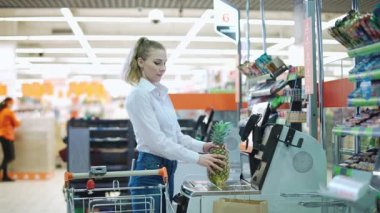 Genç bir kadın modern bir süpermarketten sebze ve meyve satın alır, bir mağazadan temas kurmadan ürün satın alır, self-servis kasası ve NFC kullanarak satın alma süreci.