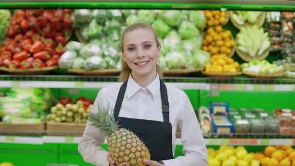 Portretul unei tinere femei zâmbitoare care lucrează într-un magazin organic, un vânzător de legume și fructe ține un ananas în mâini și se uită la camera foto . — Videoclip de stoc