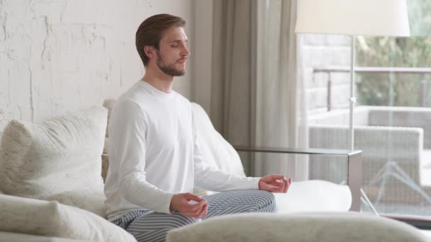 Wellness, ung man sitter på en soffa i vardagsrummet och mediterar, ett lugnt humör, koppla av i ett vitt rum fyllt med ljus, mindfulness. — Stockvideo