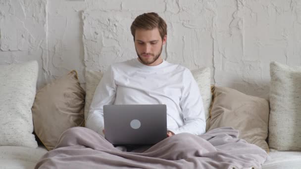 Allegro uomo caucasico si siede a casa sul divano e lavora con un computer portatile, direttore finanziario su un lavoro a distanza durante l'auto-isolamento in quarantena, un uomo fa un buon affare, emozioni positive. — Video Stock