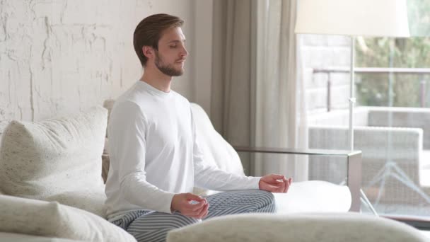 Mindfulness, joven se sienta en un sofá en la sala de estar y medita, un estado de ánimo tranquilo, relajarse en una habitación blanca llena de luz. — Vídeos de Stock