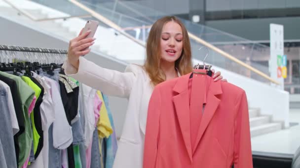 Female presents a new collection of casual clothes in a store, young woman takes selfie video of herself on smartphone, streaming video in the social network. — Stock Video