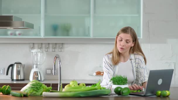 Vejetaryenlik, kadın mutfakta salata hazırlar, sağlıklı bir içecek yapmak için bitkiler ve yeşil sebzeler keser, internette dizüstü bilgisayar kullanarak kahvaltı hazırlar.. — Stok video