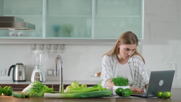 Vejetaryenlik, genç bir kadın mutfakta salata hazırlar, sağlıklı bir içecek yapmak için bitkiler ve yeşil sebzeler keser, internette dizüstü bilgisayar kullanarak kahvaltı hazırlar.. — Stok video
