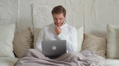 Neşeli beyaz adam evinde kanepede oturur ve dizüstü bilgisayar kullanır. Karantinada kendini izole etme, internette sörf yapma ve olumlu duygular içinde çalışan bir adam..