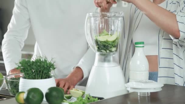 Вегетаріанство, молода пара готує коктейлі на кухні, чоловік і жінка кладуть зелені овочі інгредієнти в блендері . — стокове відео