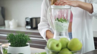 Genç dişi mutfakta meyve püresi hazırlar, yeşil sebze malzemelerini koyar ve miksere koyar..