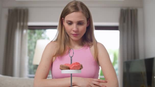 Ritratto di una giovane donna seduta in cucina a guardare biscotti maccheroni, senza osare mangiare un dolce dolce, assunzione limitata di zucchero, femmina seguendo una dieta. — Video Stock