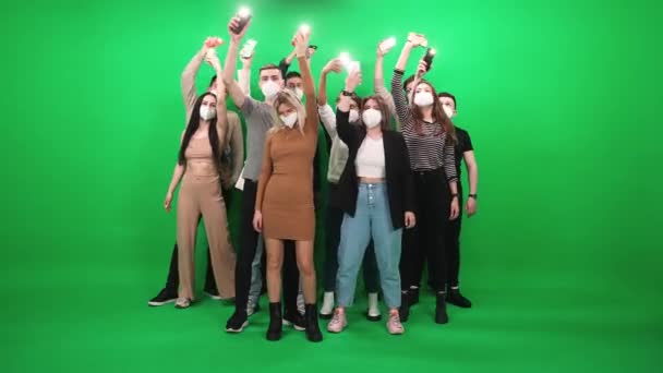 Crowd of people of different ages dancing at a music concert, people in protective medical masks holding smartphones with flashlights on, they dance and swing their arms on a green background, event — Stock Video