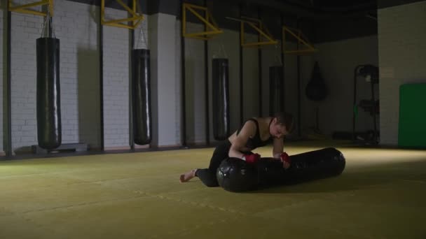Kickboxing, lutador mulher agressiva treina seus socos, bate um saco de perfuração enquanto deitado no chão, dia de treinamento no ginásio de boxe, corpo de ajuste de força, a menina bate rápido. — Vídeo de Stock