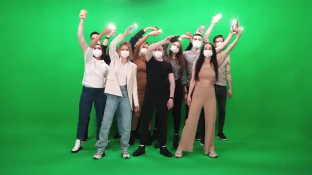 Dav lidí různého věku tančí na hudebním koncertě, lidé v ochranných lékařských maskách drží smartphony s rozsvícenými baterkami, tančí a máchají rukama na zeleném pozadí, událost — Stock video