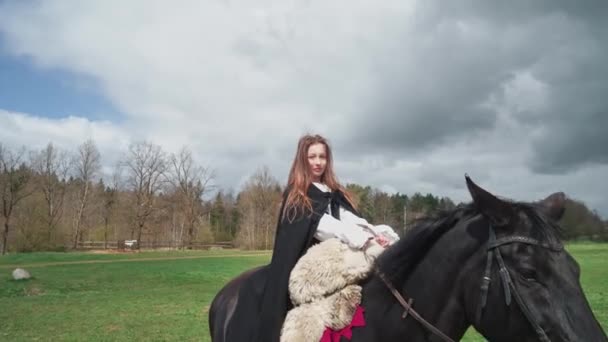 Escena medieval, hermosa hembra en un vestido medieval montando un caballo a través de un campo verde, la chica mira a la cámara, 4k cámara lenta. — Vídeo de stock