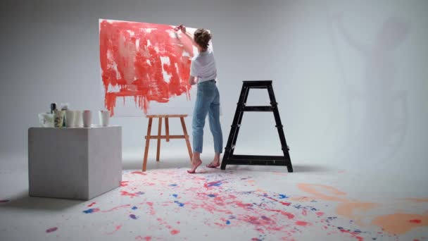 Πίσω όψη, γυναίκα καλλιτέχνης ζωγραφίζει με ένα πινέλο σε ένα μεγάλο καμβά σε ένα λευκό δωμάτιο, ένας ταλαντούχος καλλιτέχνης σχεδιάζει ένα κόκκινο χρώμα αφαίρεσης, σύγχρονη τέχνη στη ζωγραφική πετρελαίου. — Αρχείο Βίντεο