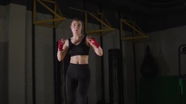 Female fighter trains his kicks, training in the boxing gym, young woman looks at the camera and trains a series of punches, 4k slow motion.