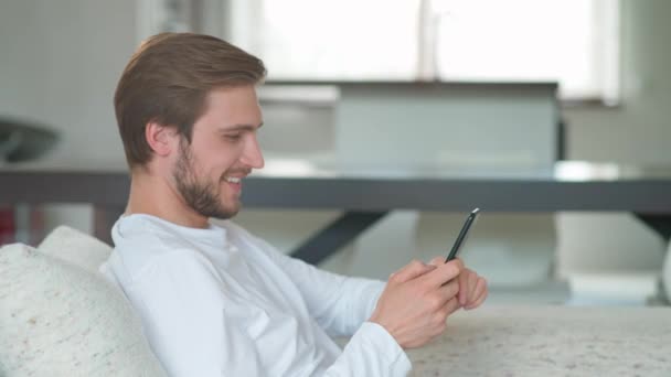 Wesoły biały mężczyzna siedzi w domu na kanapie i używa smartfona, surfuje po Internecie, spędza czas podczas samoizolacji w kwarantannie. — Wideo stockowe