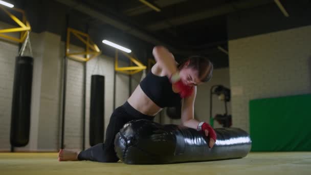 Mulher lutadora treina seus socos, bate um saco de perfuração enquanto deitado no chão, dia de treinamento no ginásio de boxe, corpo de ajuste de força, câmera lenta 4k. — Vídeo de Stock