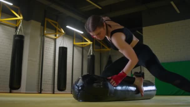 Mulher lutadora treina seus socos, bate um saco de perfuração enquanto deitado no chão, dia de treinamento no ginásio de boxe, corpo de ajuste de força, câmera lenta 4k. — Vídeo de Stock