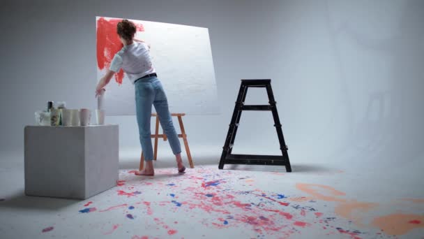 Γυναίκα καλλιτέχνης ζωγραφίζει με ένα πινέλο σε ένα μεγάλο καμβά σε ένα λευκό δωμάτιο, ένας ταλαντούχος καλλιτέχνης σχεδιάζει ένα κόκκινο χρώμα αφαίρεσης, σύγχρονη τέχνη στη ζωγραφική πετρελαίου. — Αρχείο Βίντεο