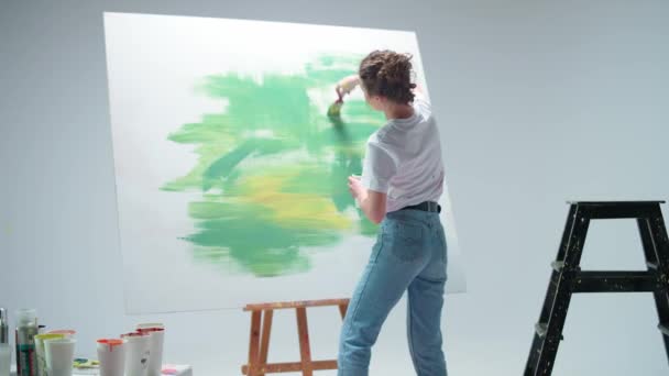 Γυναίκα καλλιτέχνης ζωγραφίζει με ένα πινέλο σε ένα μεγάλο καμβά σε ένα λευκό δωμάτιο, ένας ταλαντούχος καλλιτέχνης σχεδιάζει ένα χρώμα πράσινο αφαίρεση, σύγχρονη τέχνη στη ζωγραφική πετρελαίου. — Αρχείο Βίντεο