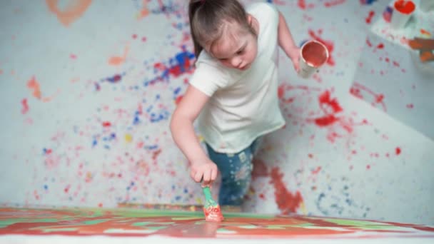 Enfant fille avec le syndrome du duvet dessine avec un pinceau sur une grande toile dans une chambre blanche, enfant fille avec des besoins spéciaux dessine une abstraction couleur rouge-bleu, 4k ralenti. — Video