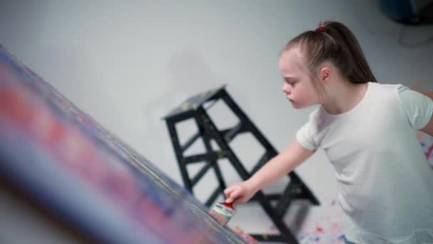Niña con síndrome de Down dibuja con un cepillo en un lienzo grande en una habitación blanca, niña con necesidades especiales dibuja un color rojo-azul abstracción, 4k cámara lenta. — Vídeo de stock