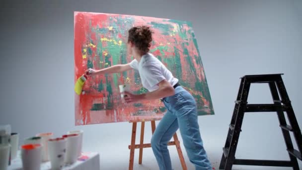 Handheld, jovem artista feminina desenha com um pincel em uma tela grande em uma sala branca, um artista talentoso desenha uma abstração de cor, arte moderna em pintura a óleo, 4k câmera lenta. — Vídeo de Stock