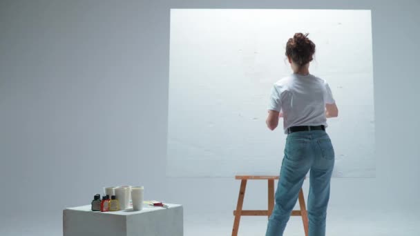 Πίσω όψη, γυναίκα καλλιτέχνης εφαρμόζει ένα στρώμα από λευκό αστάρι σε ένα μεγάλο καμβά σε ένα λευκό δωμάτιο, ένας ταλαντούχος καλλιτέχνης ετοιμάζεται να σχεδιάσετε. — Αρχείο Βίντεο