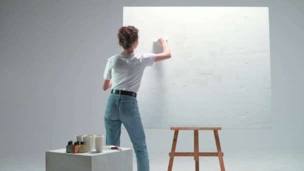 Młoda artystka nakłada warstwę białego podkładu na duże płótno w białym pomieszczeniu, utalentowana artystka przygotowująca się do rysowania. — Wideo stockowe