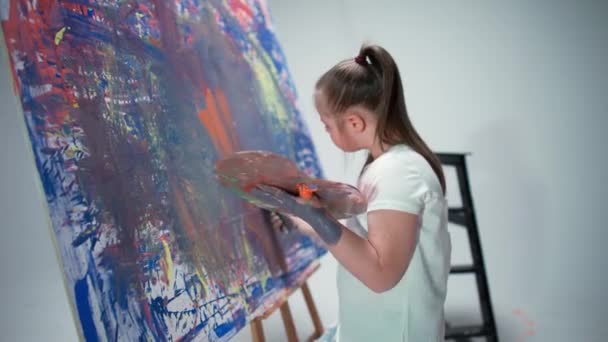 Girl with special needs draw with a brush on a large canvas in the white room, kid girl with down syndrome draw a color red-blue abstraction, 4k slow motion. — Stock video