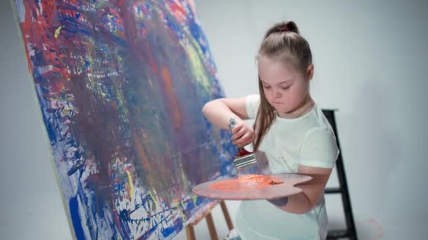 Fille avec des besoins spéciaux dessine avec un pinceau sur une grande toile dans une chambre blanche, fille enfant avec le syndrome du duvet dessine une abstraction couleur rouge-bleu, 4k au ralenti. — Video