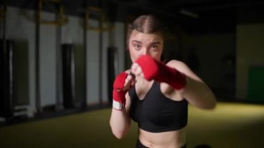 Kadın dövüşçü yumruklarını eğitiyor, boks salonunda antrenman yapıyor, genç kadın kameraya bakıyor ve bir dizi yumruk eğitiyor, ön görüş..