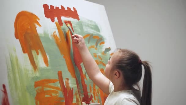 Fetița cu sindrom Down desenează cu o perie pe o pânză mare într-o cameră albă, fata cu nevoi speciale atrage o abstracție de culoare roșu-albastru . — Videoclip de stoc